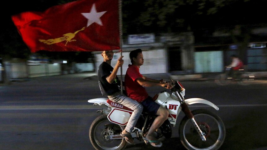 Two supporters of the National League for Democracy ride a motorbike at night while carrying a big flag.