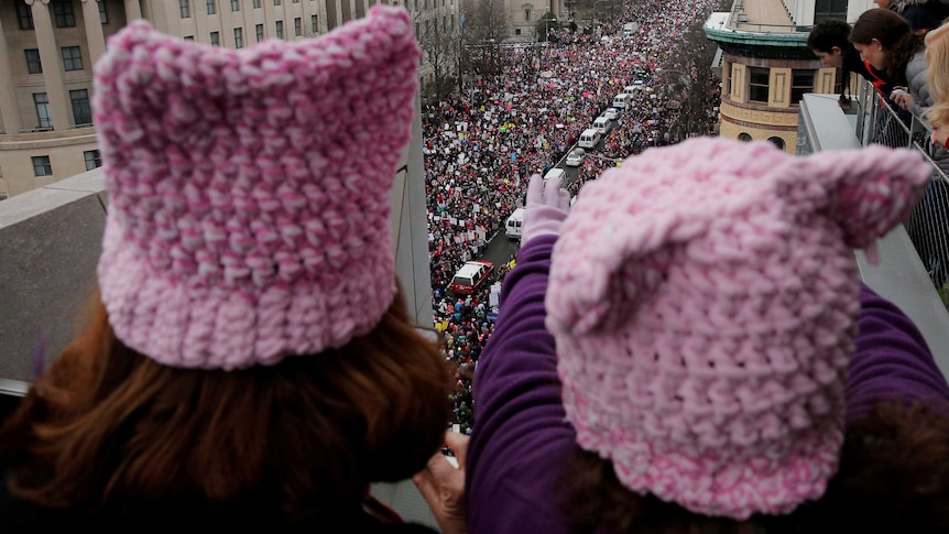 A close-up photo of two women from behind wearing pink beanies. There is a large crowd marching in the background.