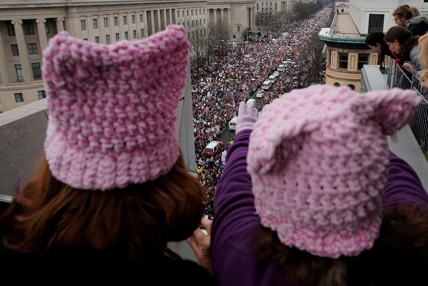 A close-up photo of two women from behind wearing pink beanies. There is a large crowd marching in the background.