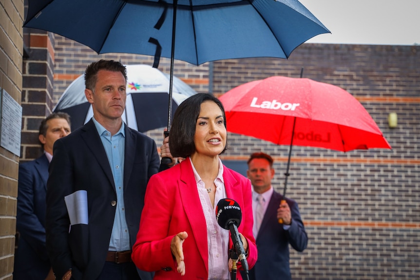 a woman talking into microphones while standing outdoors as a man stands behind holding an umbrella