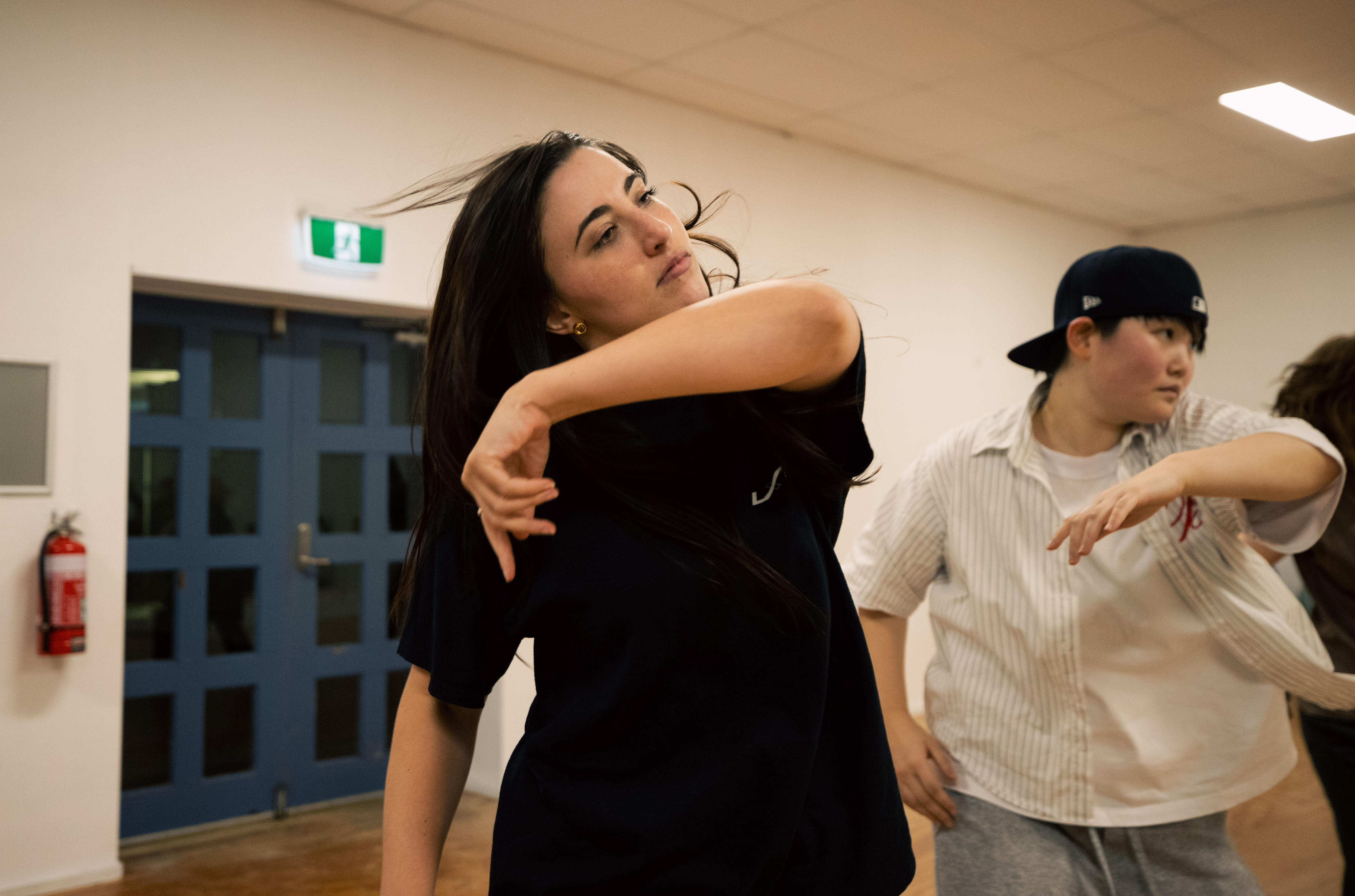 Una mujer de cabello oscuro y camiseta oscura baila con otras personas en una habitación.