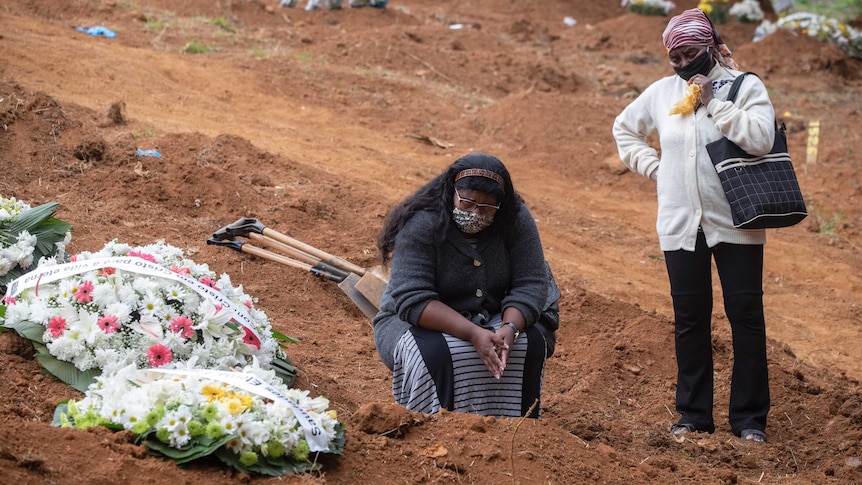 Two women crouch and stand by a grave with flowers on it in a dirt ceremony