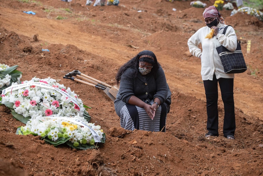 Two women crouch and stand by a grave with flowers on it in a dirt ceremony