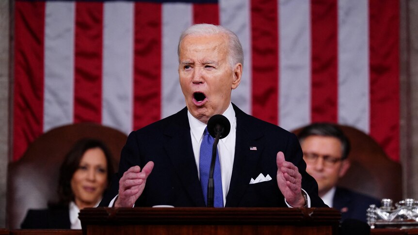 Joe Biden wearing a suit and blue tie stands in front of a US flag speaking into a microphone.