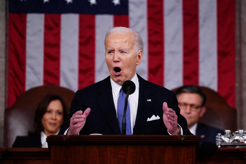 Joe Biden wearing a suit and blue tie stands in front of a US flag speaking into a microphone.