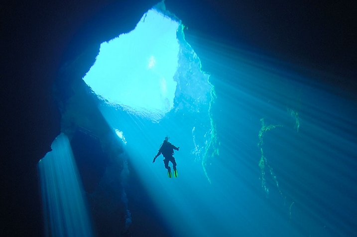 A lone diver swims up towards the opening of a cave. Light shines through from above, illuminating dark corners of the cavern