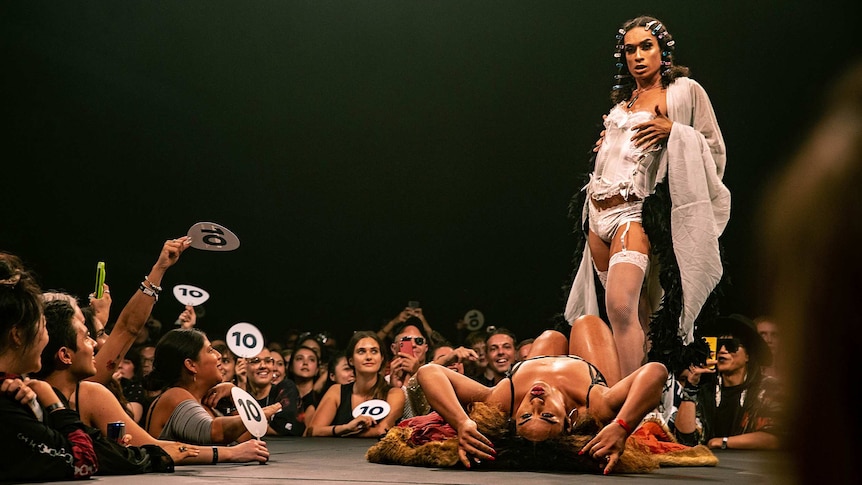 Colour photo of Kilia Tipa lying on back next to a standing dancer on stage at Sissy Ball 2019.