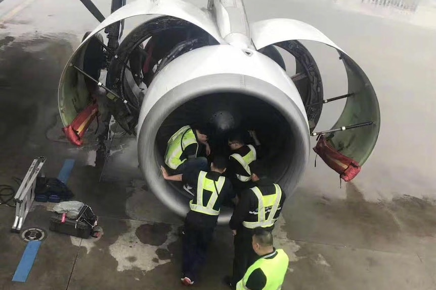 Five plane mechanics gather around a plane engine as they work to retrieve a coin thrown by a passenger.