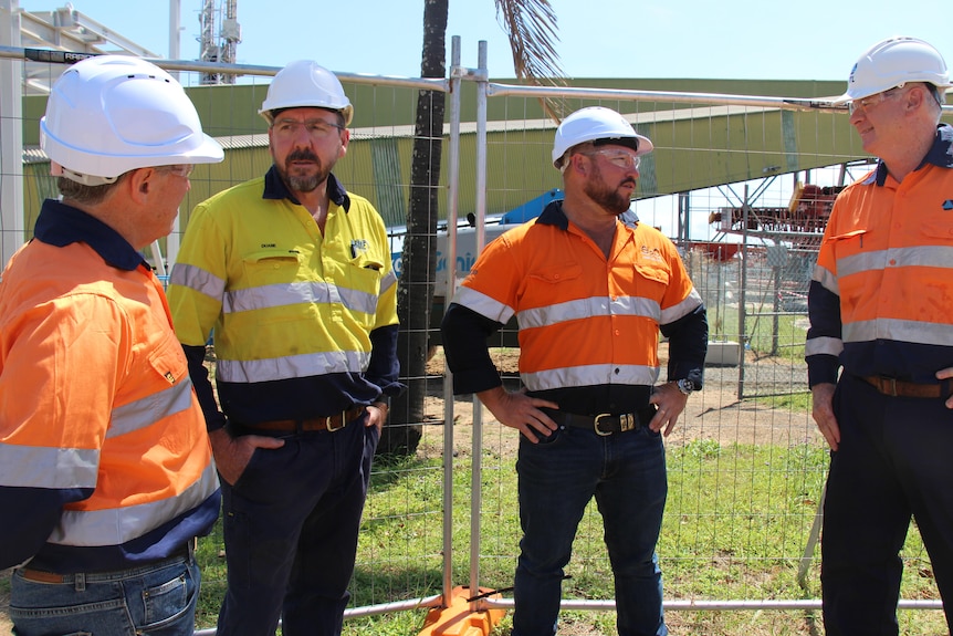 Four men in high-vis uniform shirts and hard hats stand with their hands on their hips, infrastructure scaffolding behind