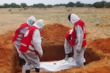 A group of four people dressed in white PPE and wearing red vests stand around an unmarked grave.