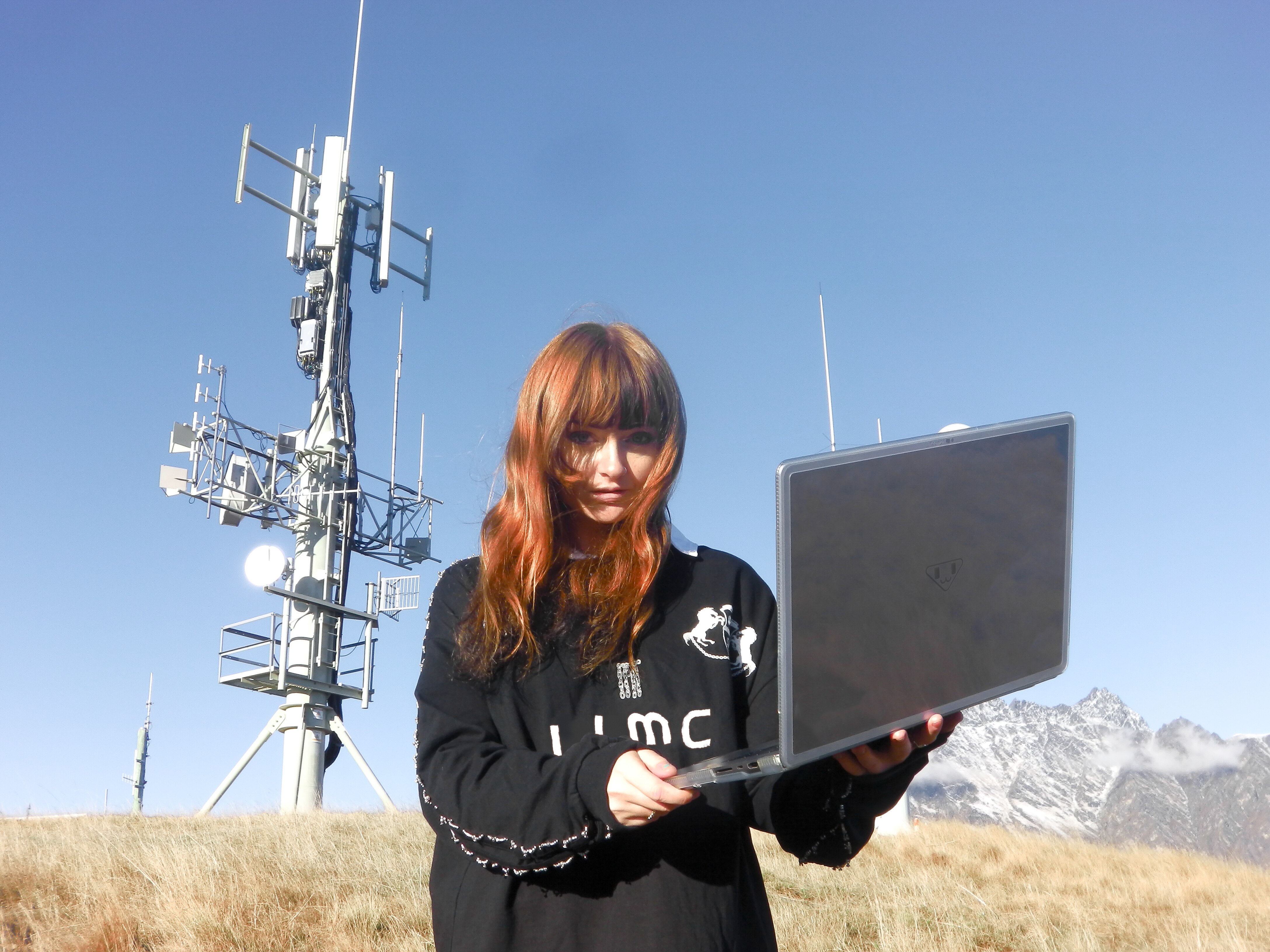 Ninajirachi wears a black long sleeve shirt and stands in a field in front of a satellite pole with her computer