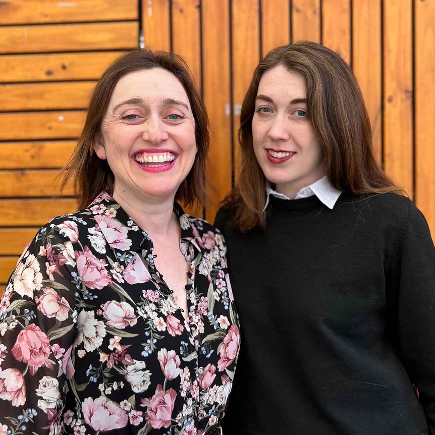 Two women standing close smiling in front of a wood lined wall