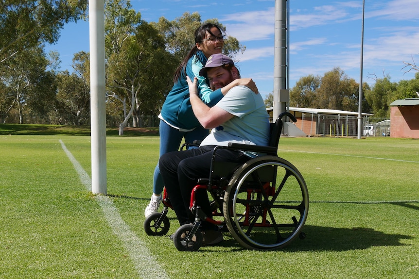 A man in a wheelchair hugs a woman on underneath the goal posts on a football field.