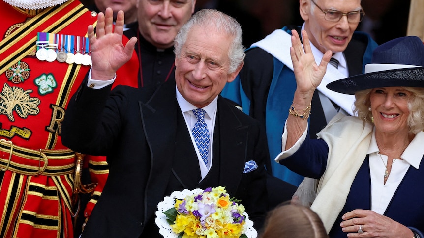 King Charles III stands on steps next to Queen Consort Camilla holding a bouquet of flowers. 