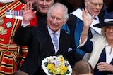 King Charles III stands on steps next to Queen Consort Camilla holding a bouquet of flowers. 