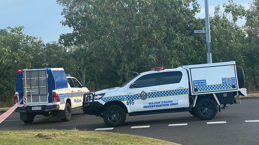 An officer walks through a park, with police tape and a police car in the foreground.