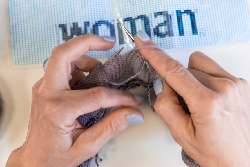 Kate Just's hands knitting, a knitting pattern with the word 'woman' underneath her hands