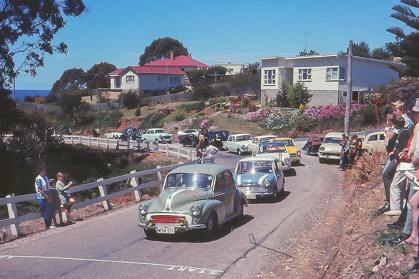 Cars roll to the the start line on the winding road lined with houses.