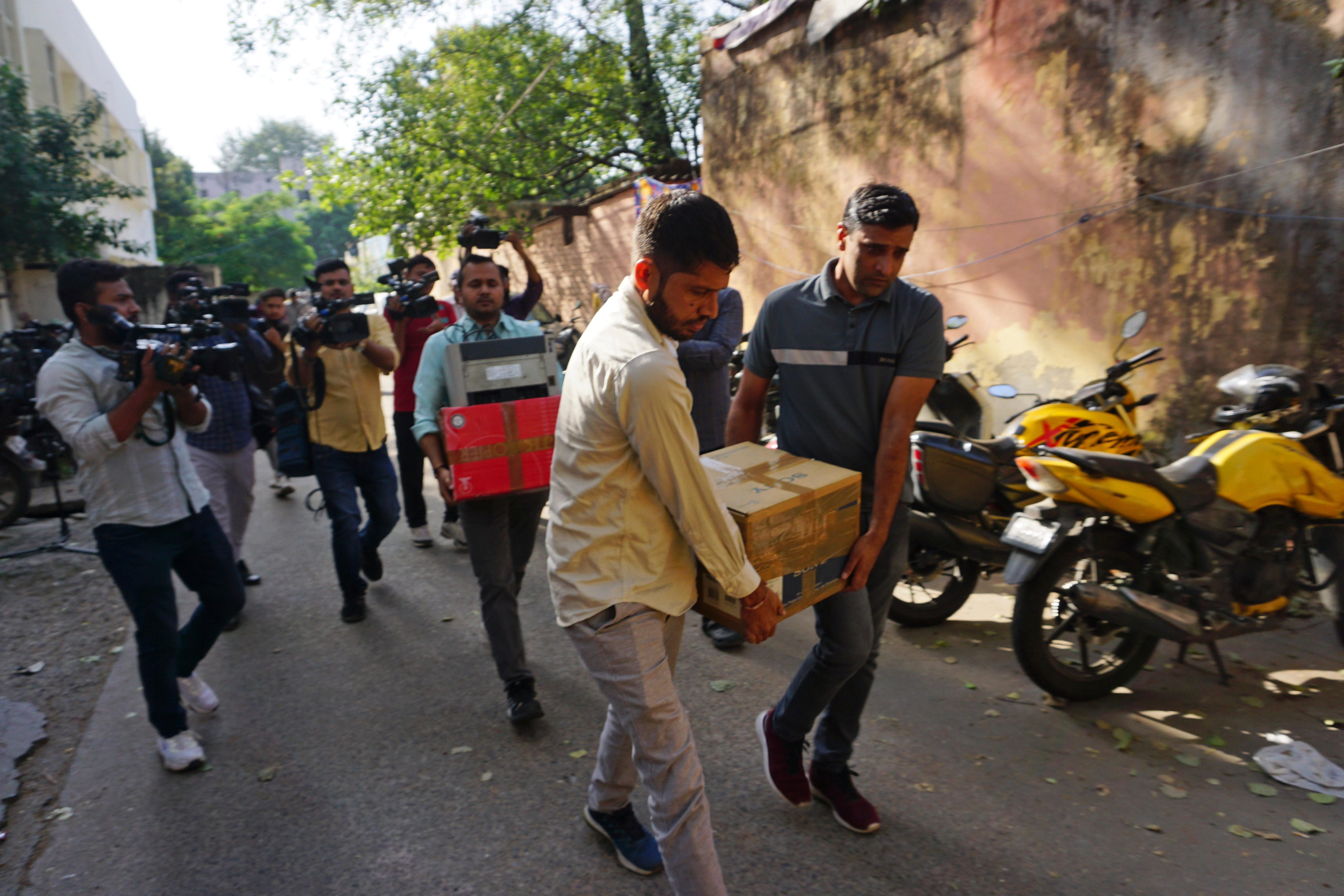 Three men walk down a street carrying boxes, followed by other men filming them with video cameras.