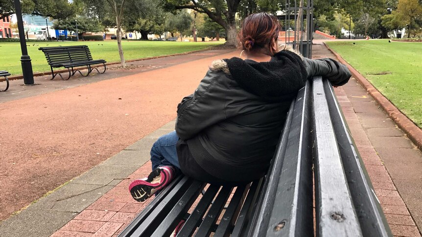 A woman sits on a bench in a park looking away from the camera, so you cannot see her face.