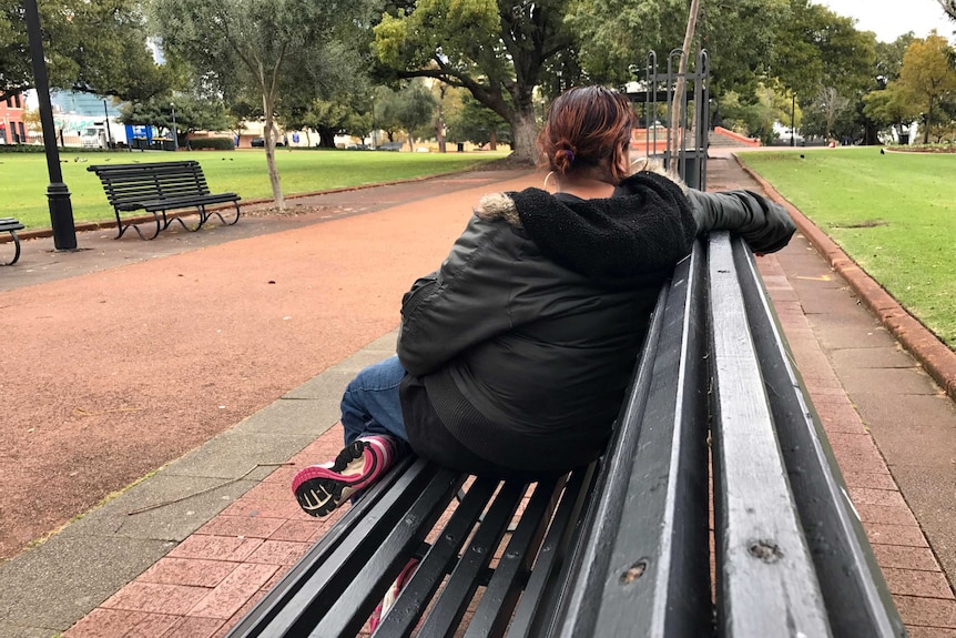 A woman sits on a bench in a park looking away from the camera, so you cannot see her face.