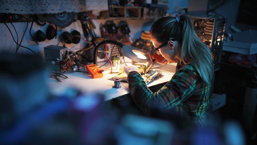 A woman sits at a desk with a small light, creating something