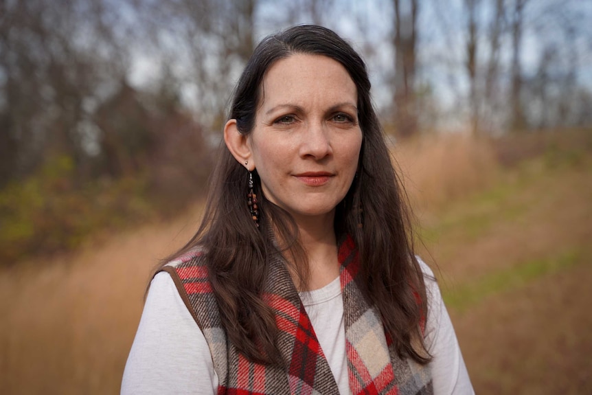 a woman with long hair in a tartan vest looks impassively at the camera