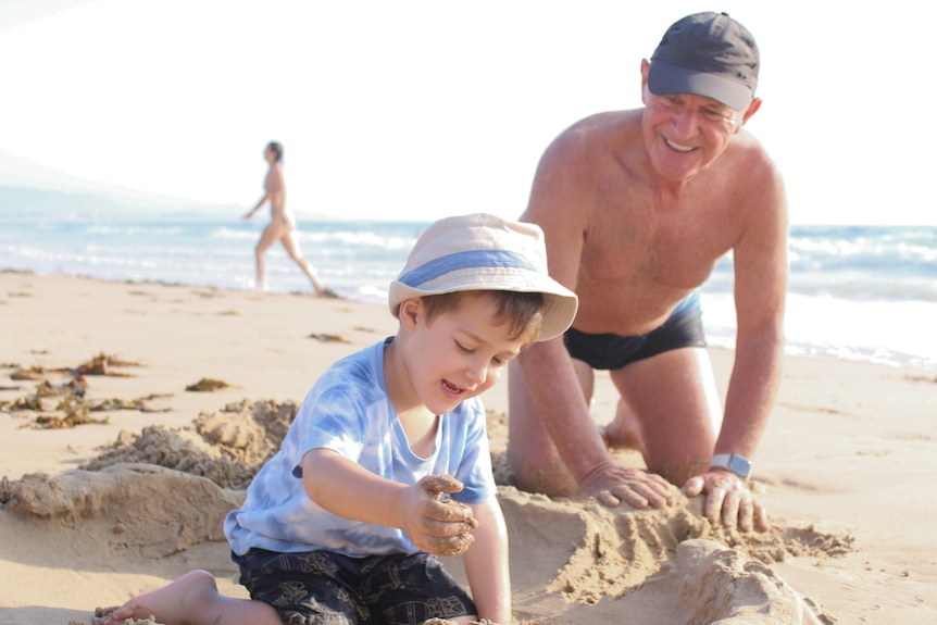 Young boy wearing hat and blue shirt, playing in sand, shirtless grandfather smiling, watches on