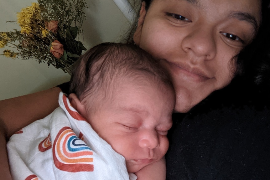 A young woman lays on a bed with a tiny baby snuggled into her chest. The baby is sleeping peacefully, the woman smiles