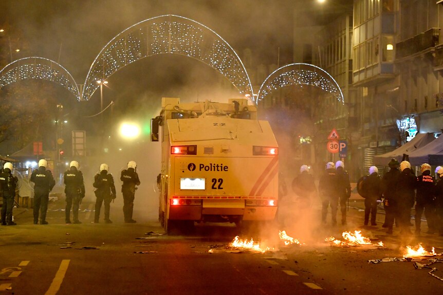 police line up either side of a water cannon in Brussels at night where small fires burn on the road