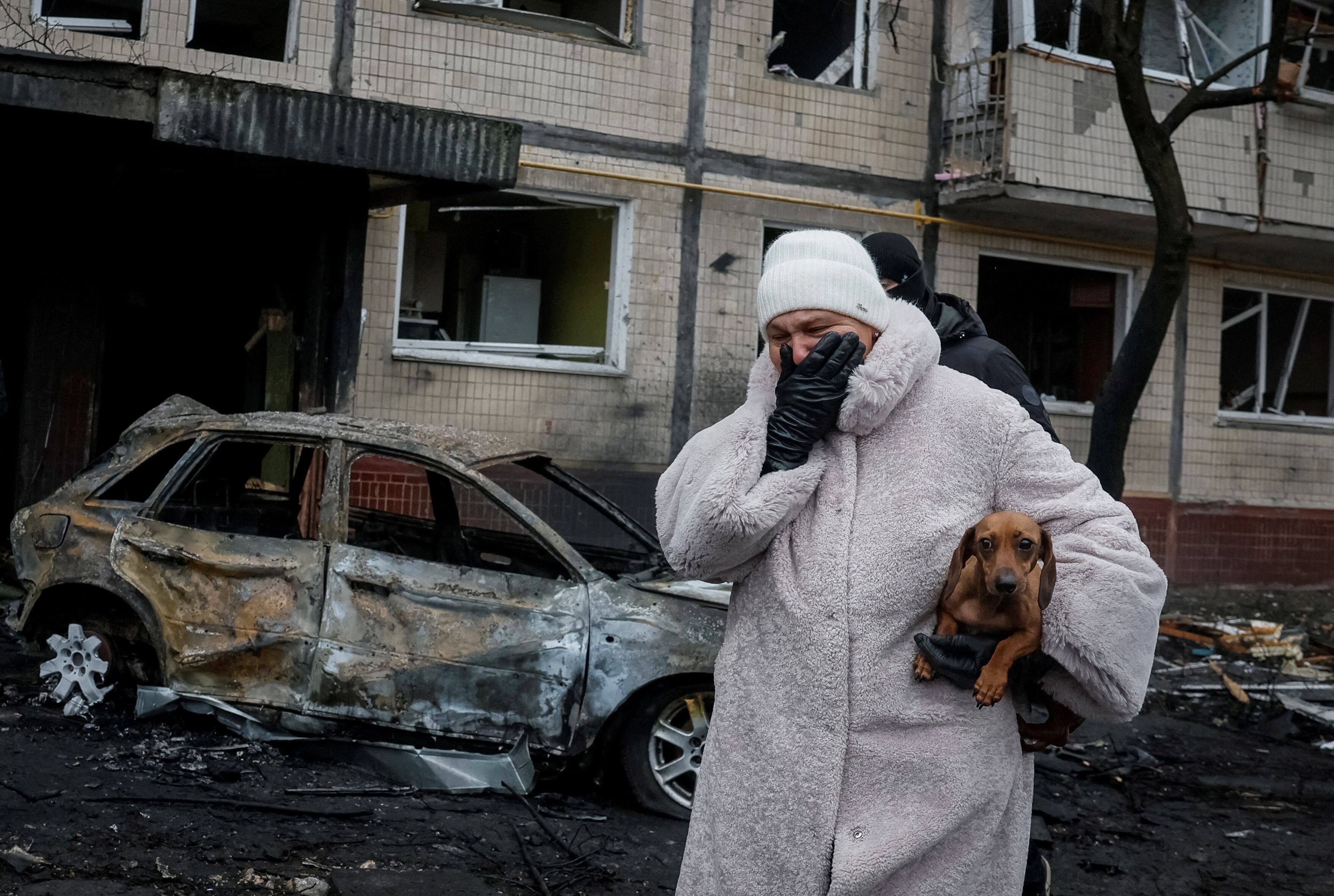 A woman in a long coat, beanie, gloves, holding a dog, crying, next to a damaged building and burned car.
