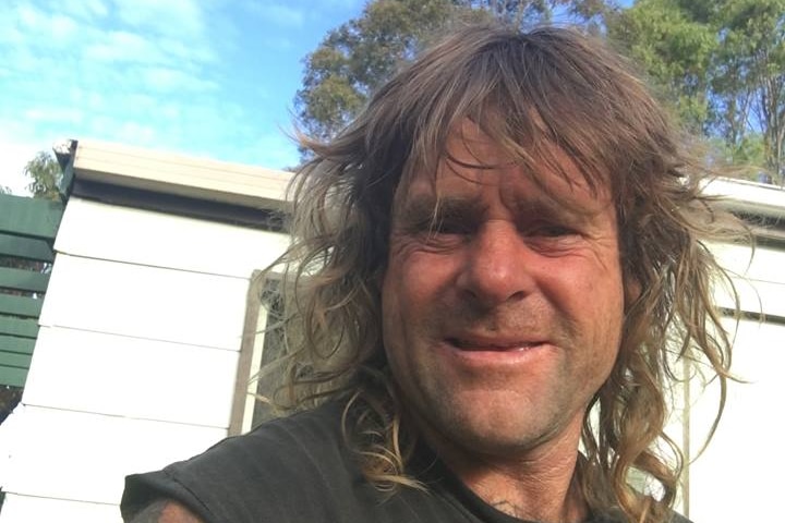 A man with scruffy hair squints while taking a selfie in bushland.