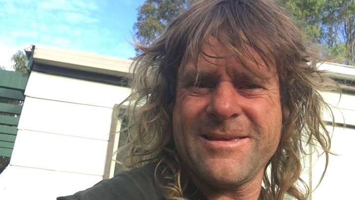 A man with scruffy hair squints while taking a selfie in bushland.