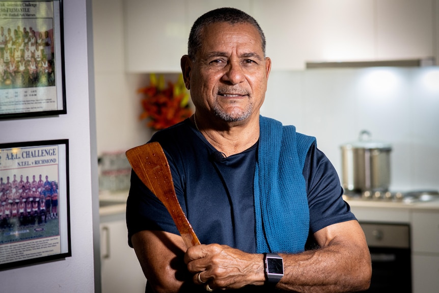 A man wearing a blue t shirt holds a wooden spoon and smiles to the camera. A tea towel is over his shoulder.