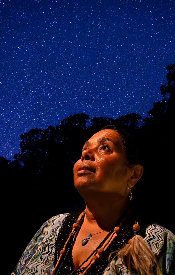 Close up of Indigenous woman looking up into the sky, silhouetted by dark bushes and blue sky with stars 