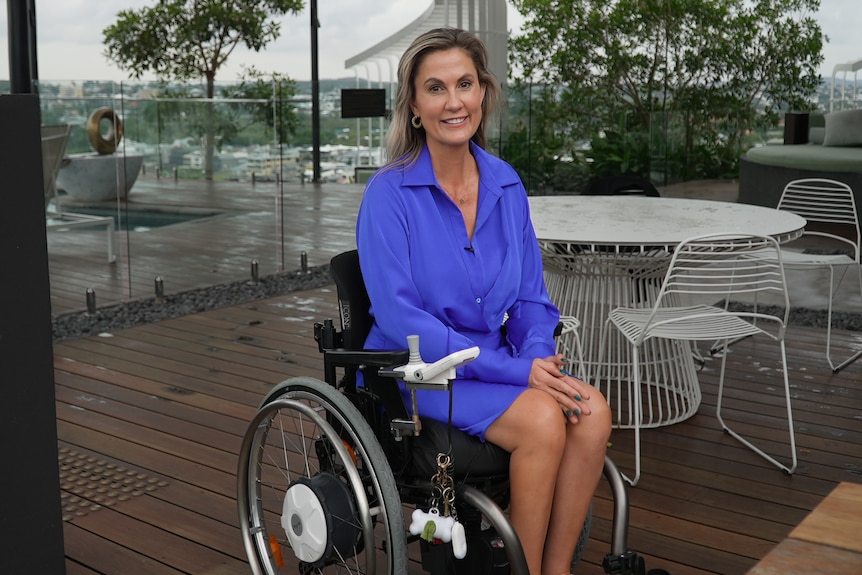 Karni Liddell sits in a powerchair on a wooden deck with a table behind her. She wears a blue dress and blue nail polish.