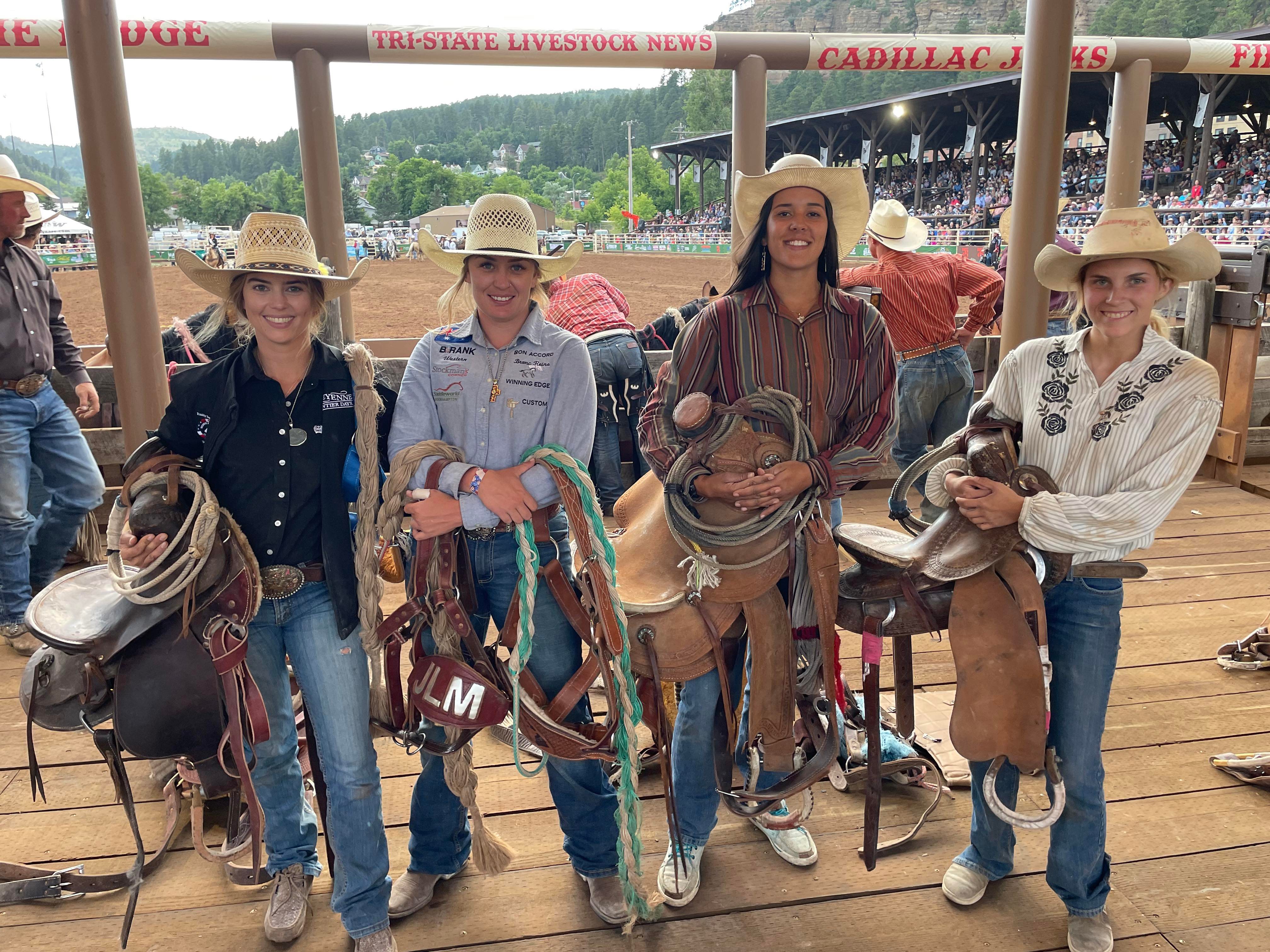 Four women carrying saddlery and ride gear stand side by side with a rodeo ground behind them.