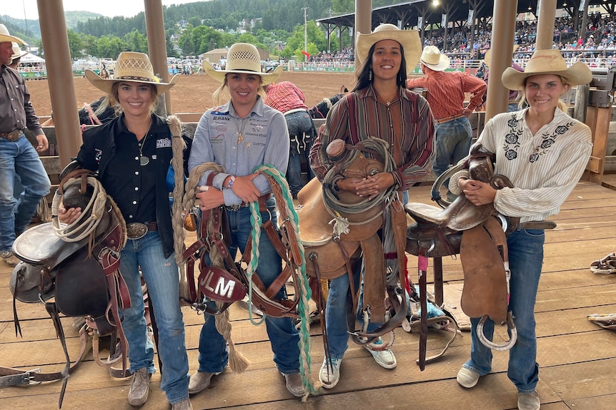 Four women carrying saddlery and ride gear stand side by side with a rodeo ground behind them.