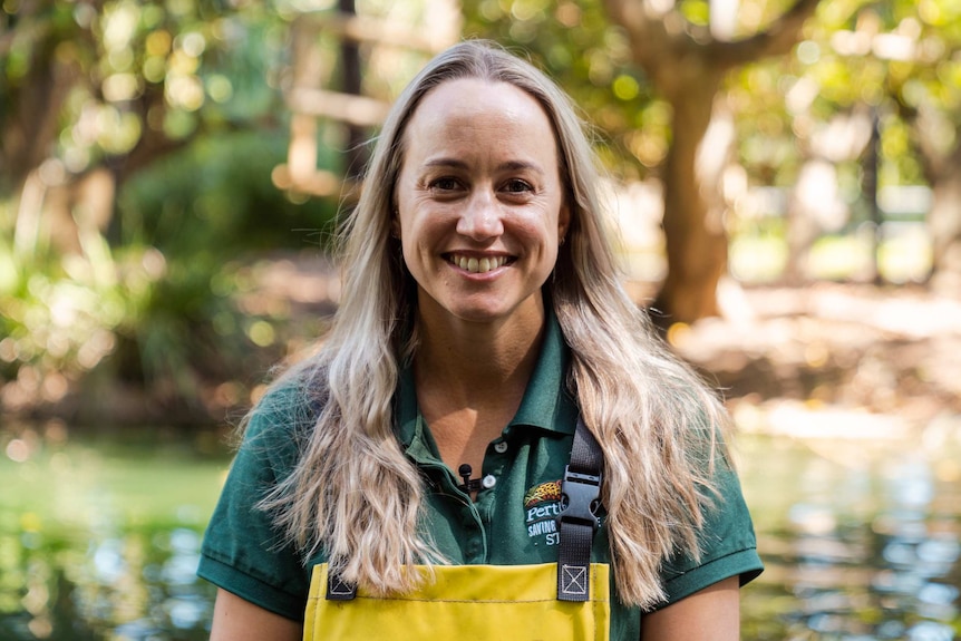A close-up of Holly Thompson wearing a dark green t-shirt and yellow overalls with trees in the background.