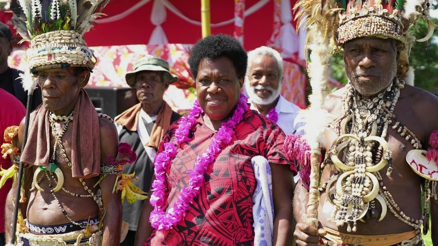 A woman in a red and black  patterned shirt stands with men in traditional Papua New Guinean dress