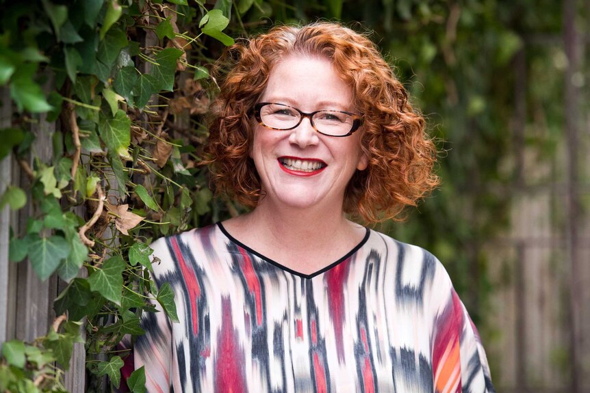 A woman with curly red hair, glasses and a brightly coloured shirt smiles widely. Behind her a green vine grows along a fence.