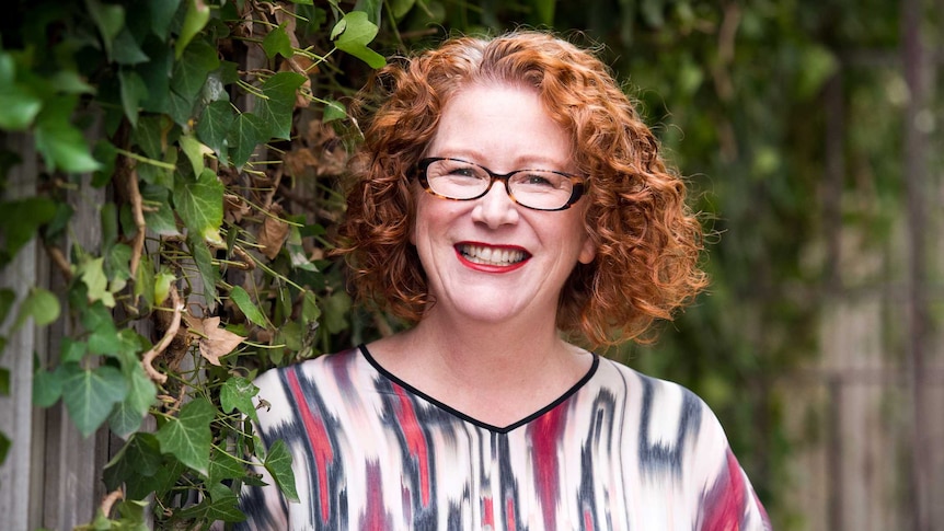 A woman with curly red hair, glasses and a brightly coloured shirt smiles widely. Behind her a green vine grows along a fence.