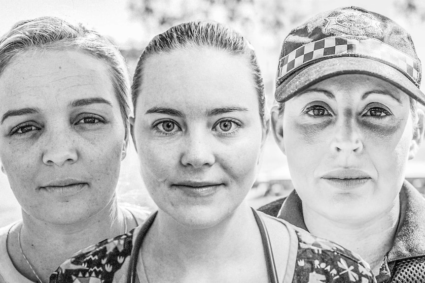 Three women stand photographed in black and white staring directly at the camera.