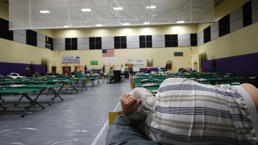 A man lies on one of countless cots set up in rows in a large building.