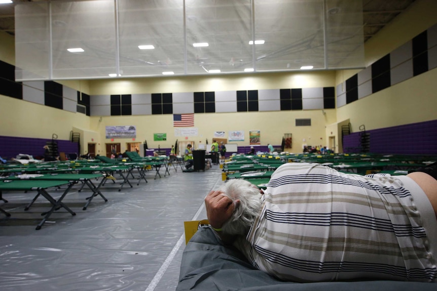 A man lies on one of countless cots set up in rows in a large building.