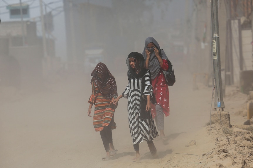Three Indian girls walk on a dusty street while covering their heads with scarves.