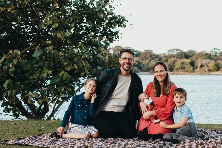 A father, mother, and three children sitting together smiling on a picnic rug.