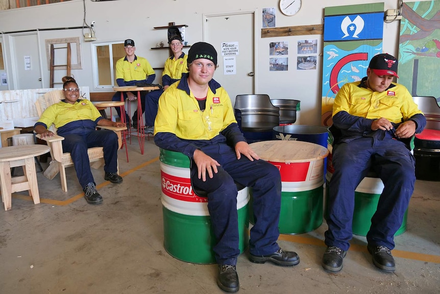 Five youths dressed in workwear sit on seats made from upcycled oil drums and wooden pallets