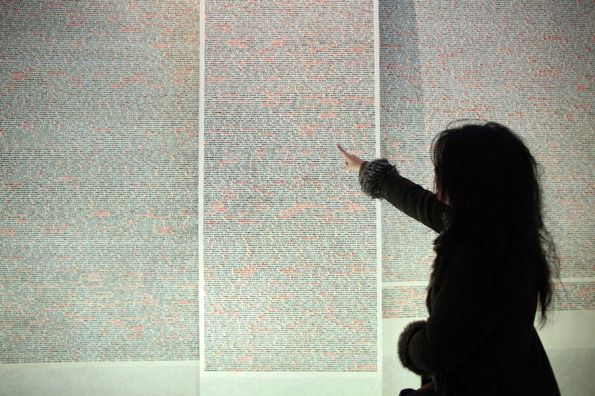 A woman points at a wall with tens of thousands of names in black and red on the wall of a synagogue.