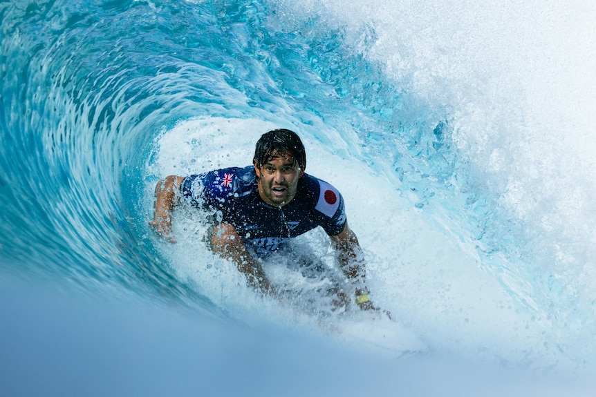 A surfer, wearing Australian and Japanese flags on his outfit, uses his hand to steady himself as he moves through a big wave.